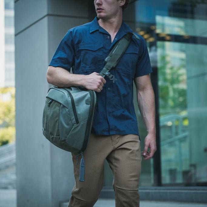 Wearing a blue short-sleeved shirt and tan pants, a person carries a Vertx Keryx Sling while walking by large glass windows in an urban setting.