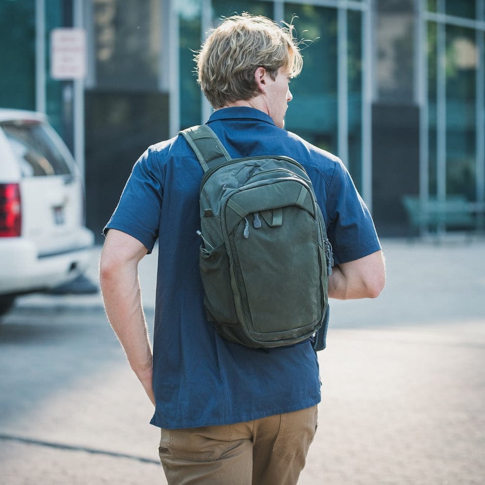 A person with blond hair walks down a paved street, carrying a green Vertx Transit Sling 3.0 Backpack featuring a Rapid Access Tab. They are dressed in a navy blue shirt and brown pants, with a white vehicle and glass building visible behind them.