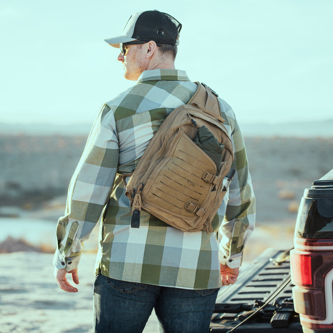 A man in a plaid shirt and black cap stands near a pickup with its tailgate down in the desert, wearing sunglasses and a tan Vertx Siege 15L Tactical Sling crossbody bag, ready for life's adventures.