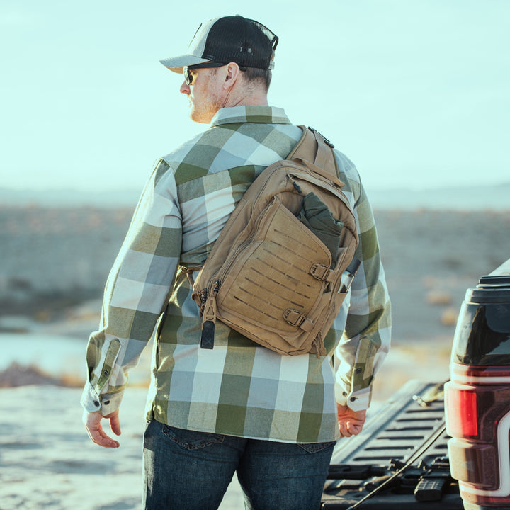A man in a plaid shirt and black cap stands near a pickup with its tailgate down in the desert, wearing sunglasses and a tan Vertx Siege 15L Tactical Sling crossbody bag, ready for life's adventures.
