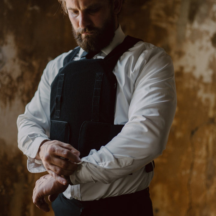 A bearded person in a white shirt and black vest, adjusting their sleeve, stands by a window in a textured room. The soft light mirrors the subtle protection offered by Premier Body Armor's Discreet Defender Bundle - Agile PE Armor without compromising style.