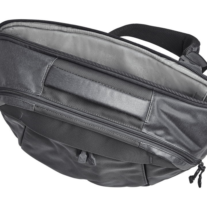 The Vertx Keryx Sling is a gray fabric backpack with a black handle, featuring two large zippered compartments for body armor inserts. Its top view highlights the closed zippers and its simple, practical design made of durable material.
