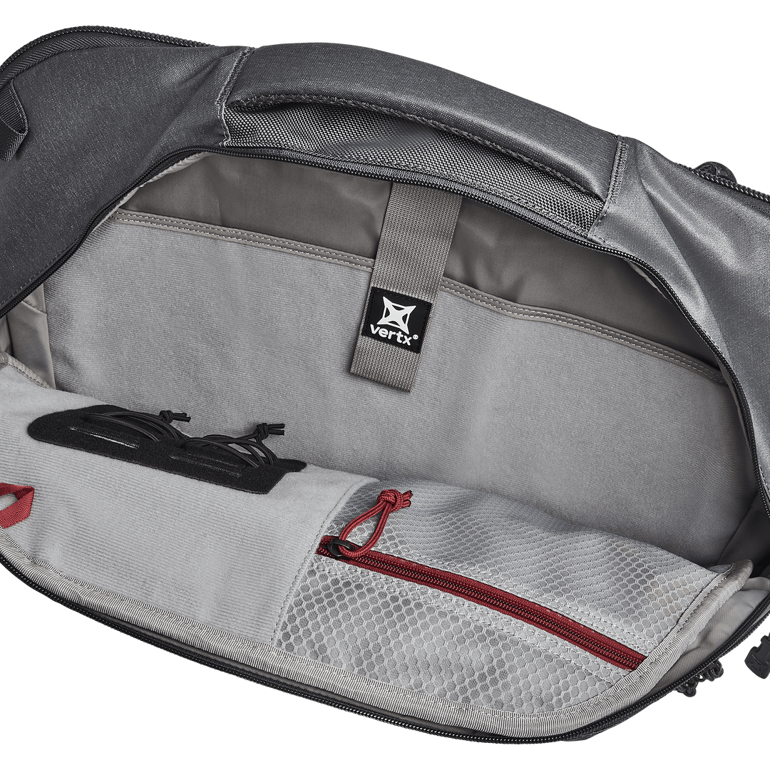 The open backpack reveals a roomy compartment ideal for EDC customization. Inside, glasses in a black case sit beside a white mesh zippered pocket with a bold red zipper. A gray "Vertx" strap suggests compatibility with both the Vertx Keryx Sling and an armor insert.