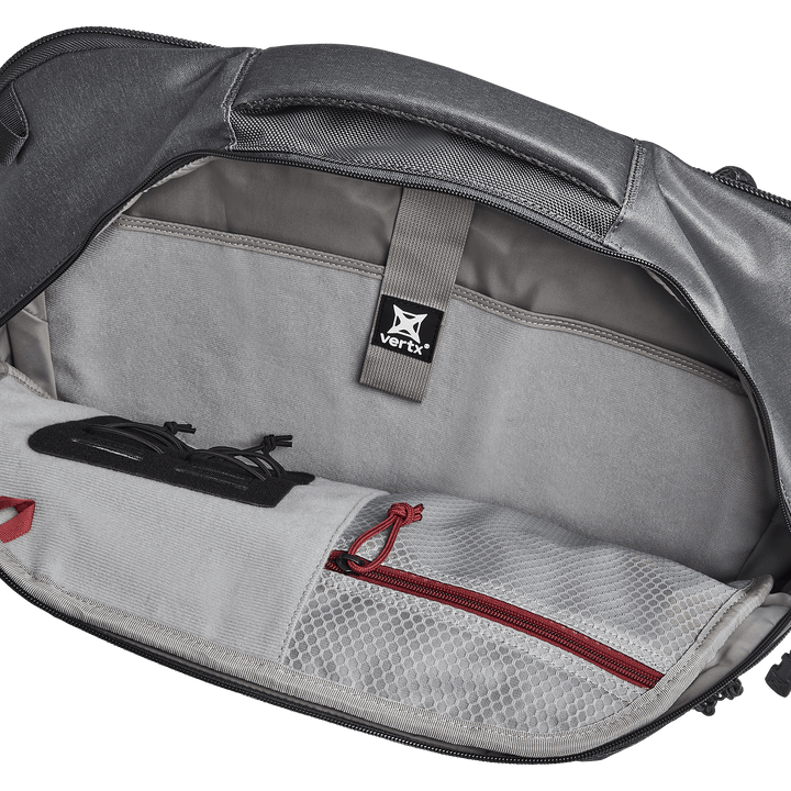 The open backpack reveals a roomy compartment ideal for EDC customization. Inside, glasses in a black case sit beside a white mesh zippered pocket with a bold red zipper. A gray "Vertx" strap suggests compatibility with both the Vertx Keryx Sling and an armor insert.