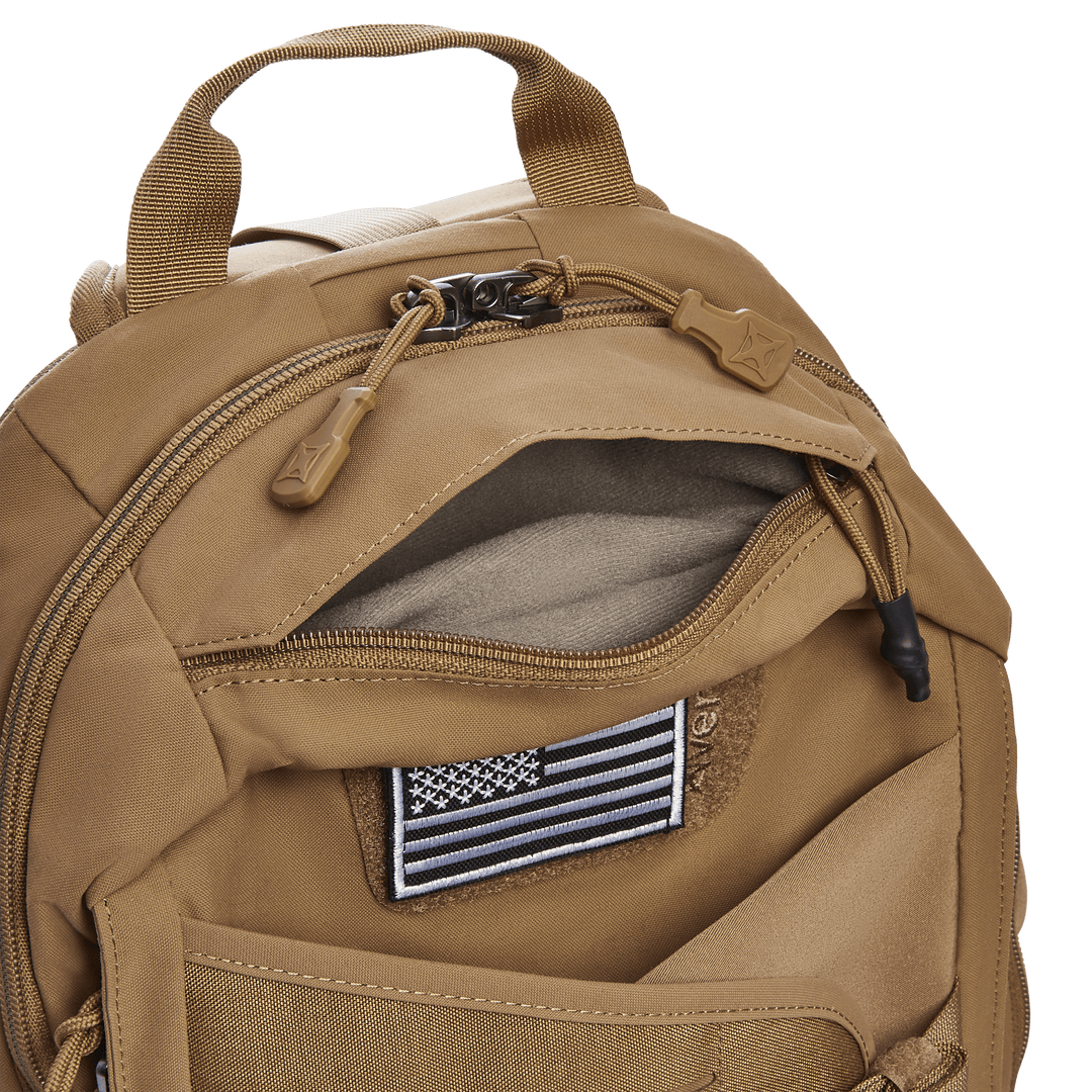 The Vertx Siege 15L Tactical Sling backpack in tan offers multiple zippered compartments, including an open one displaying a black and white American flag patch. It features a top handle, dual zippers with fabric pulls, visible stitching details, and a CCW compartment for tactical use.