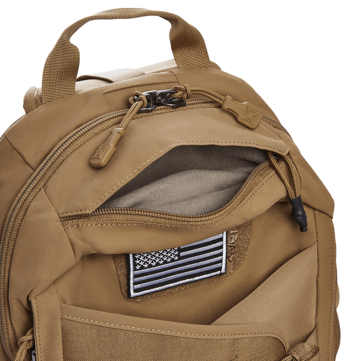 The Vertx Siege 15L Tactical Sling backpack in tan offers multiple zippered compartments, including an open one displaying a black and white American flag patch. It features a top handle, dual zippers with fabric pulls, visible stitching details, and a CCW compartment for tactical use.