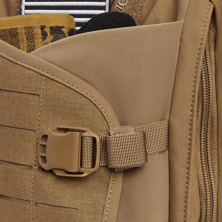 Close-up of a tan backpack pocket on the Vertx Siege 15L Tactical Sling, holding various items like a striped object and folded yellow fabric. Features include a plastic buckle, zipper, multiple stitchings, and a concealed CCW compartment for tactical readiness.