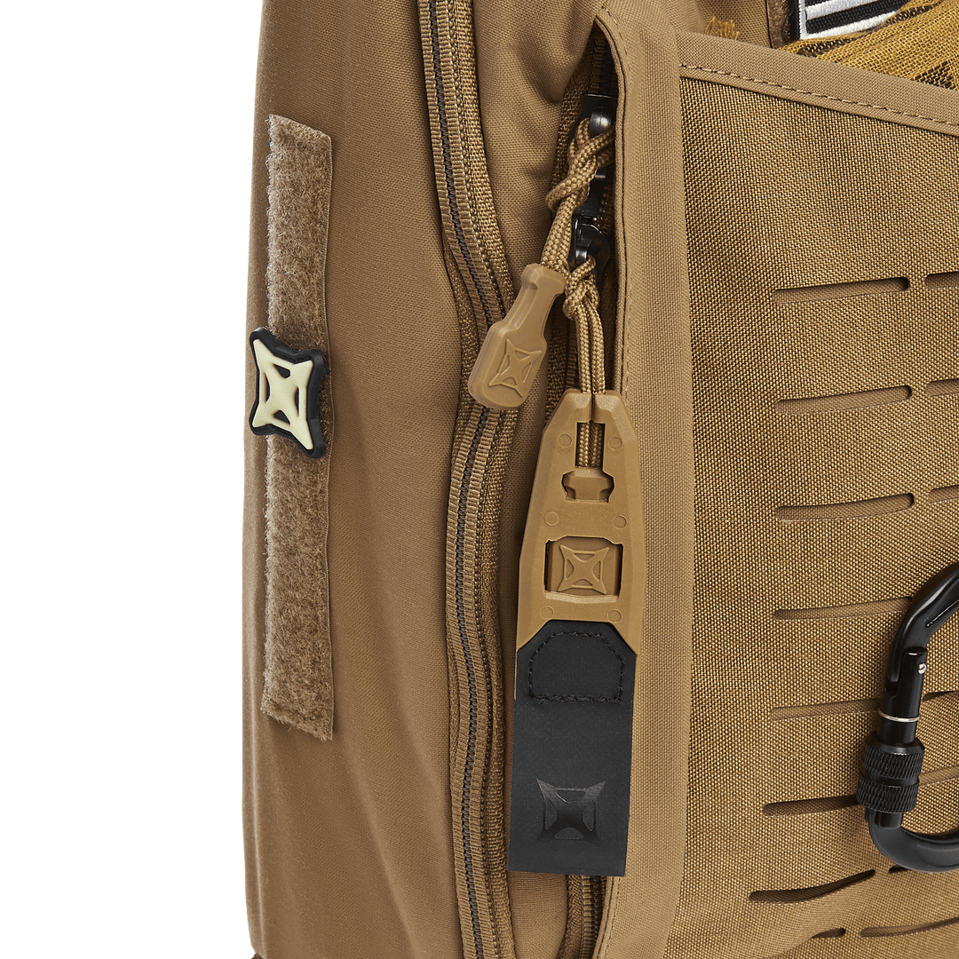 Close-up of a Vertx Siege 15L Tactical Sling highlights zipper pulls, a black and tan carabiner, and a Velcro patch on fabric with tactical horizontal slits and reinforced stitching against a solid green background.