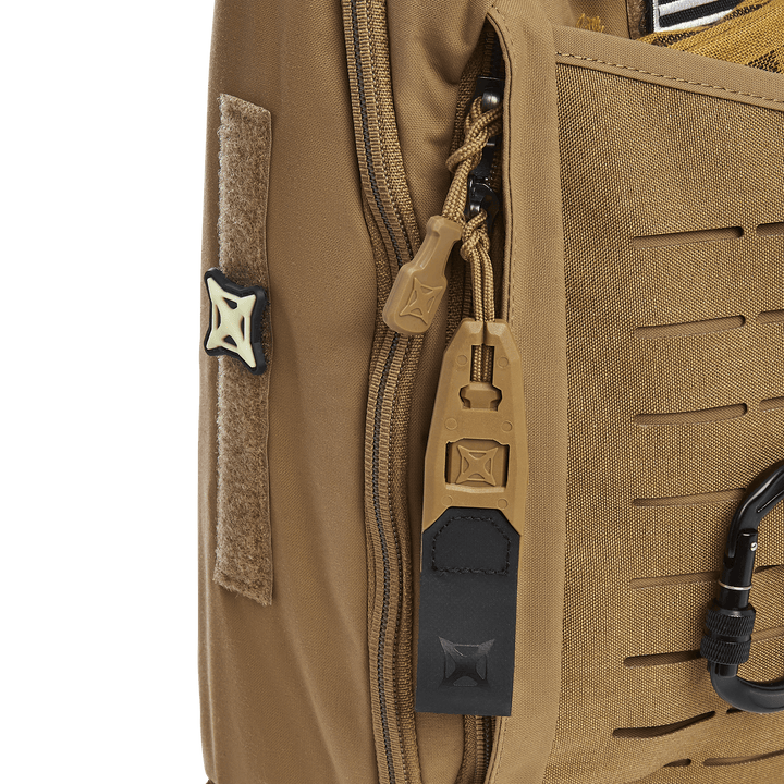 Close-up of a Vertx Siege 15L Tactical Sling highlights zipper pulls, a black and tan carabiner, and a Velcro patch on fabric with tactical horizontal slits and reinforced stitching against a solid green background.