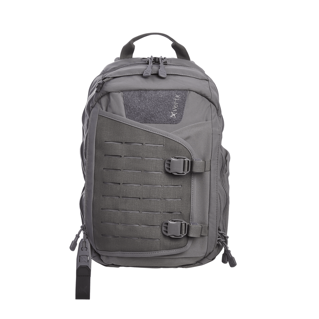 The Vertx Siege 15L Tactical Sling in Wolf Grey exudes readiness with multiple compartments, buckle closures, and a CCW compartment. It features a central zippered section, adjustable padded straps, and a top carry handle with the Vertx logo prominently displayed on the upper front panel.