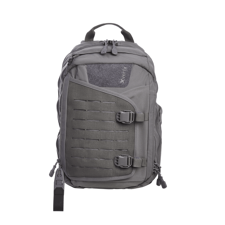 The Vertx Siege 15L Tactical Sling in Wolf Grey exudes readiness with multiple compartments, buckle closures, and a CCW compartment. It features a central zippered section, adjustable padded straps, and a top carry handle with the Vertx logo prominently displayed on the upper front panel.