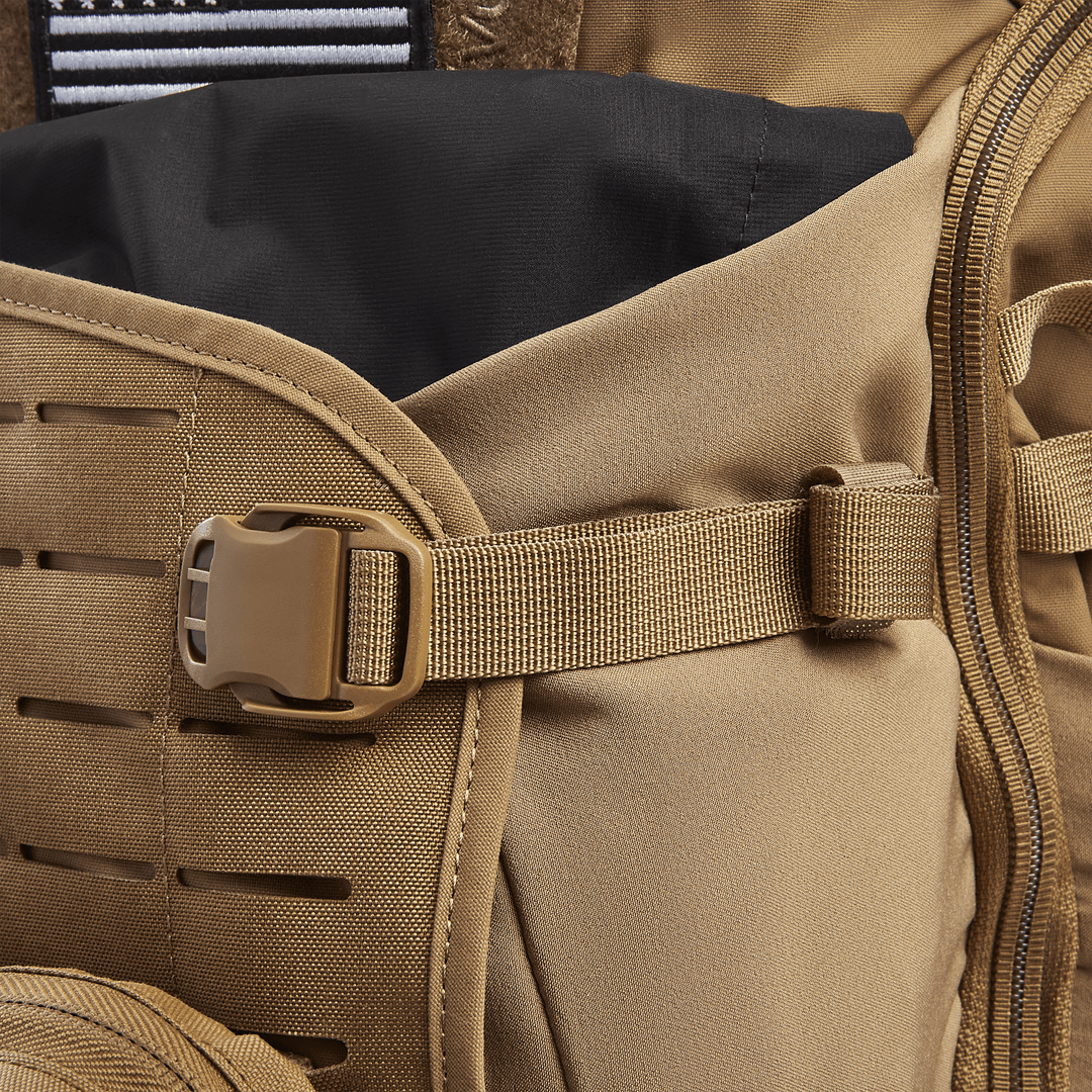 A close-up of a tan Vertx Siege 25L Tactical Pack reveals a plastic buckle joining sections, horizontal fabric slits, and a black top flap, highlighting its rugged design for tactical readiness and versatility.