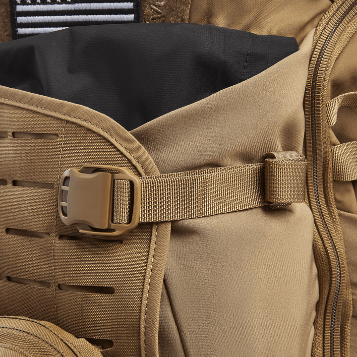 A close-up of a tan Vertx Siege 25L Tactical Pack reveals a plastic buckle joining sections, horizontal fabric slits, and a black top flap, highlighting its rugged design for tactical readiness and versatility.