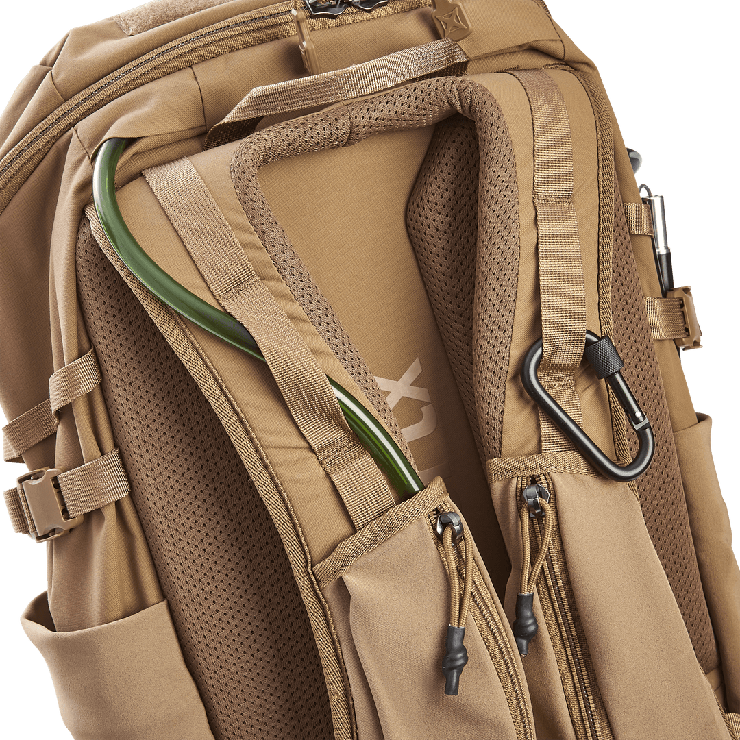 The Vertx Siege 25L Tactical Pack is a tan backpack with padded shoulder straps, featuring a hydration tube holder and black carabiner. Designed for tactical readiness, it includes multiple zippered compartments and adjustable straps.