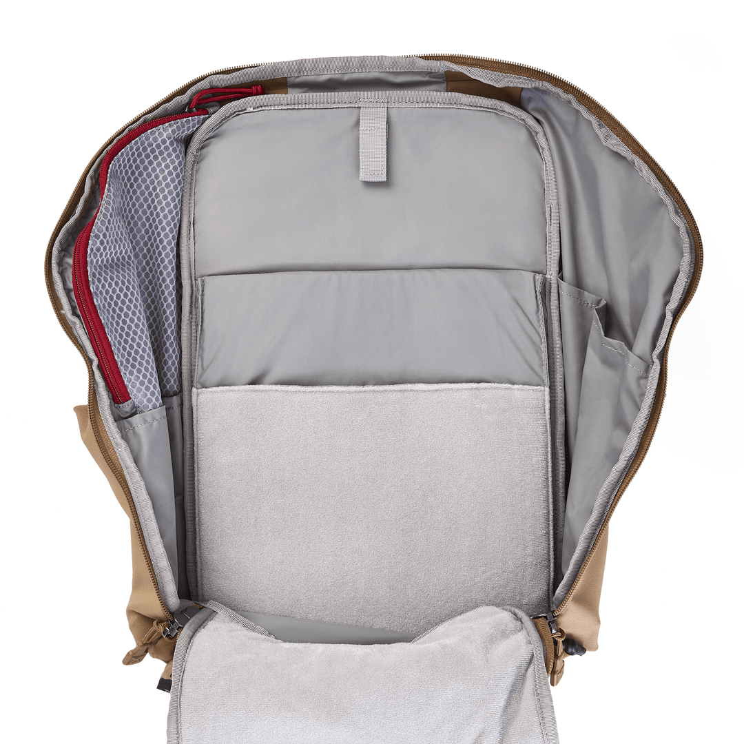 The open Vertx Siege 25L Tactical Pack showcases its tactical readiness with a brown exterior. Inside are multiple compartments, including a large padded laptop section and a mesh pocket with red lining, revealing its versatile functionality and organized layout when fully unzipped.
