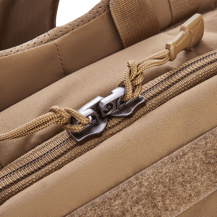 Close-up of the tan Vertx Siege 25L Tactical Pack zipper, featuring a metal double pull with paracord ties. The partially open backpack illustrates tactical readiness with its sturdy fabric, reinforced stitching, and nearby Velcro patch highlighting versatile functionality.