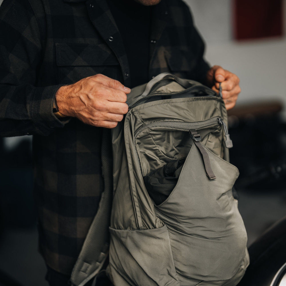 A person in a dark checkered shirt stands by a motorcycle, adjusting their gray Vertx Long Walks Pack 28L. The focus is on their hands and the backpack, with blurred motorcycle details in the foreground.