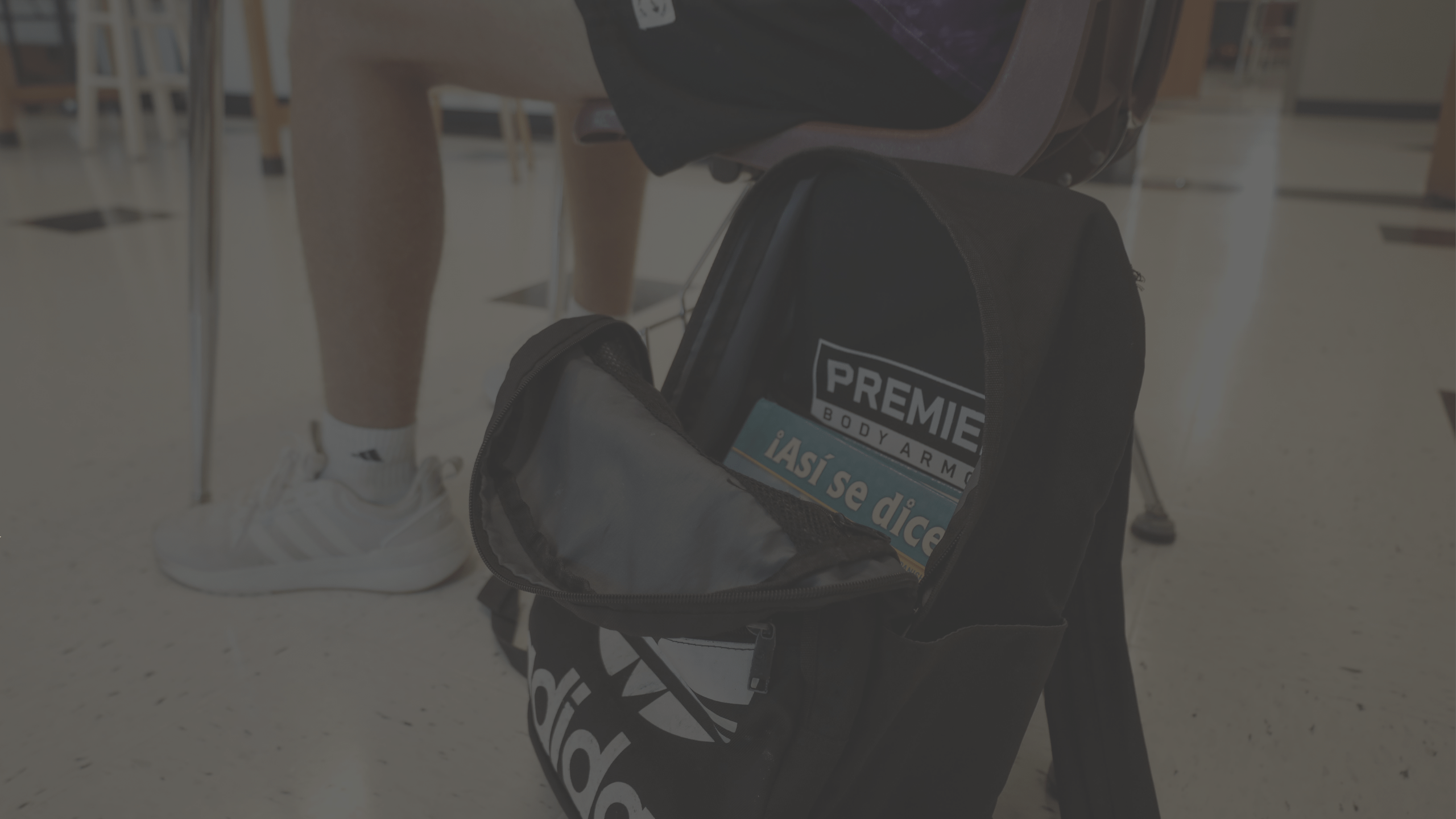 A person wearing white Adidas sneakers and black shorts sits at a desk. A black Adidas backpack on the floor has an open compartment revealing a book titled "¡Así se dice!" and a bag of Premier Body Armor inside. The scene appears to be in a classroom.