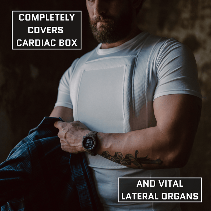 A man wears the Premier Body Armor Everyday Armor T-Shirt 360 (Armored), pulling down his plaid shirt to reveal a white vest labeled "Completely covers cardiac box and vital lateral organs.