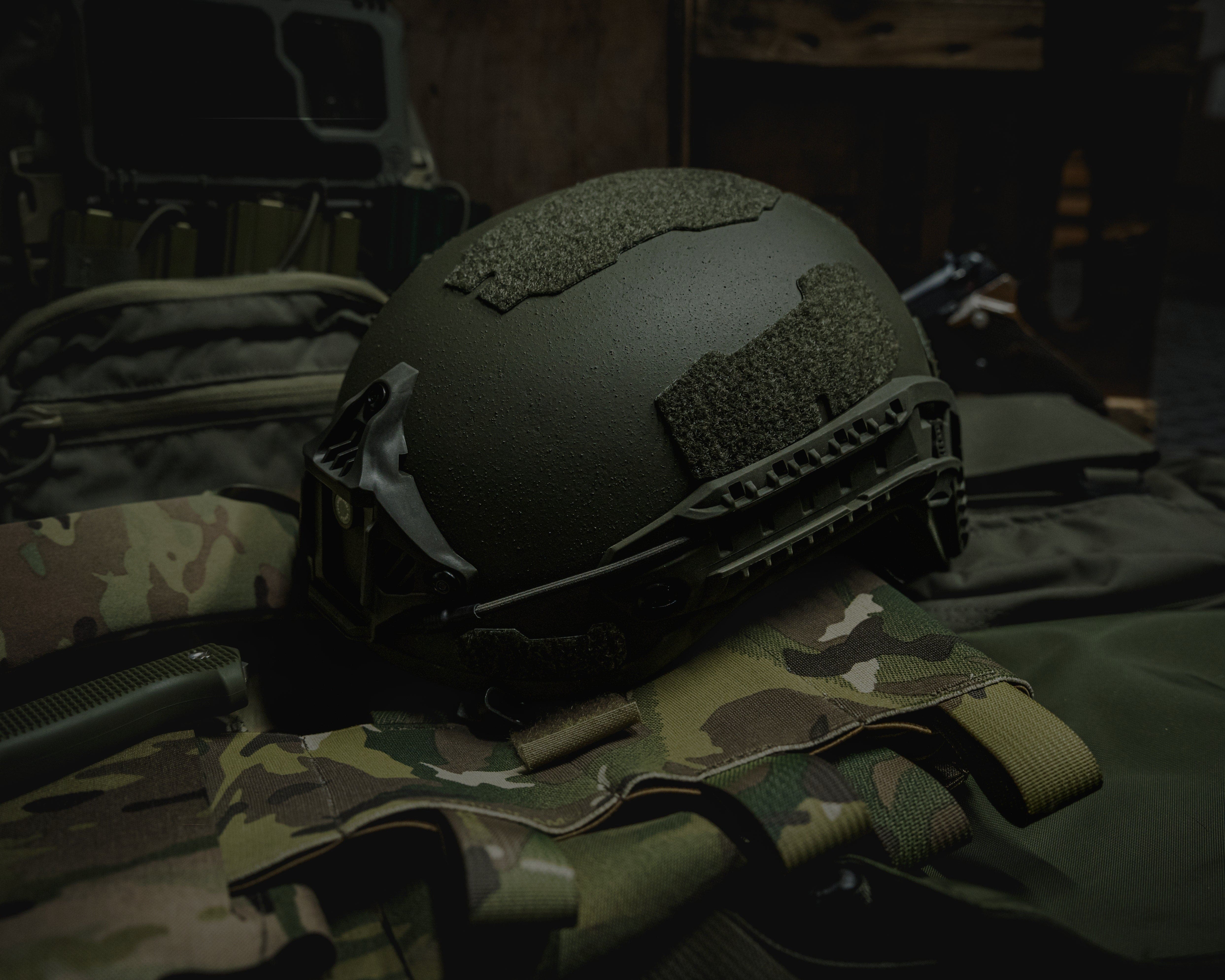 A military-style helmet with tactical attachments rests on camouflage clothing and gear, including a vest and backpack, in a dimly lit setting.