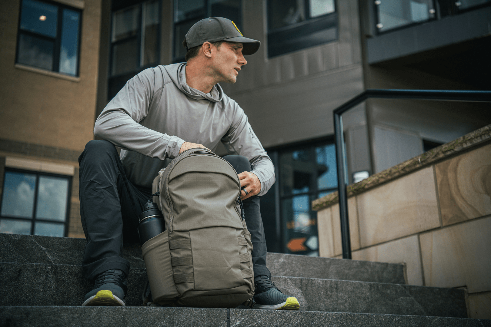 A man in a grey hoodie, dark pants, and a cap sits on outdoor steps, holding a large grey backpack. A water bottle is visible in the backpack’s side pocket. Buildings are in the background.