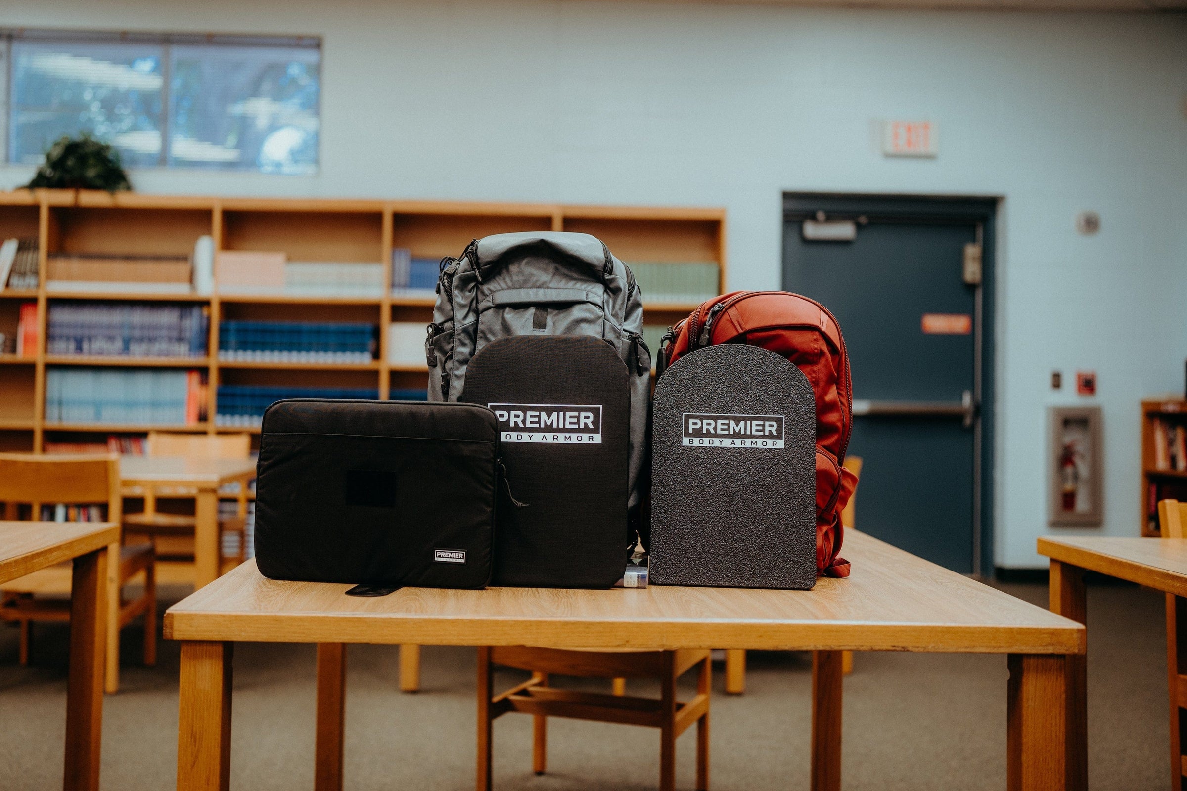 Three backpacks with Premier Body Armor labels and an additional protective panel are displayed on a wooden table in a library setting with bookshelves and chairs in the background.