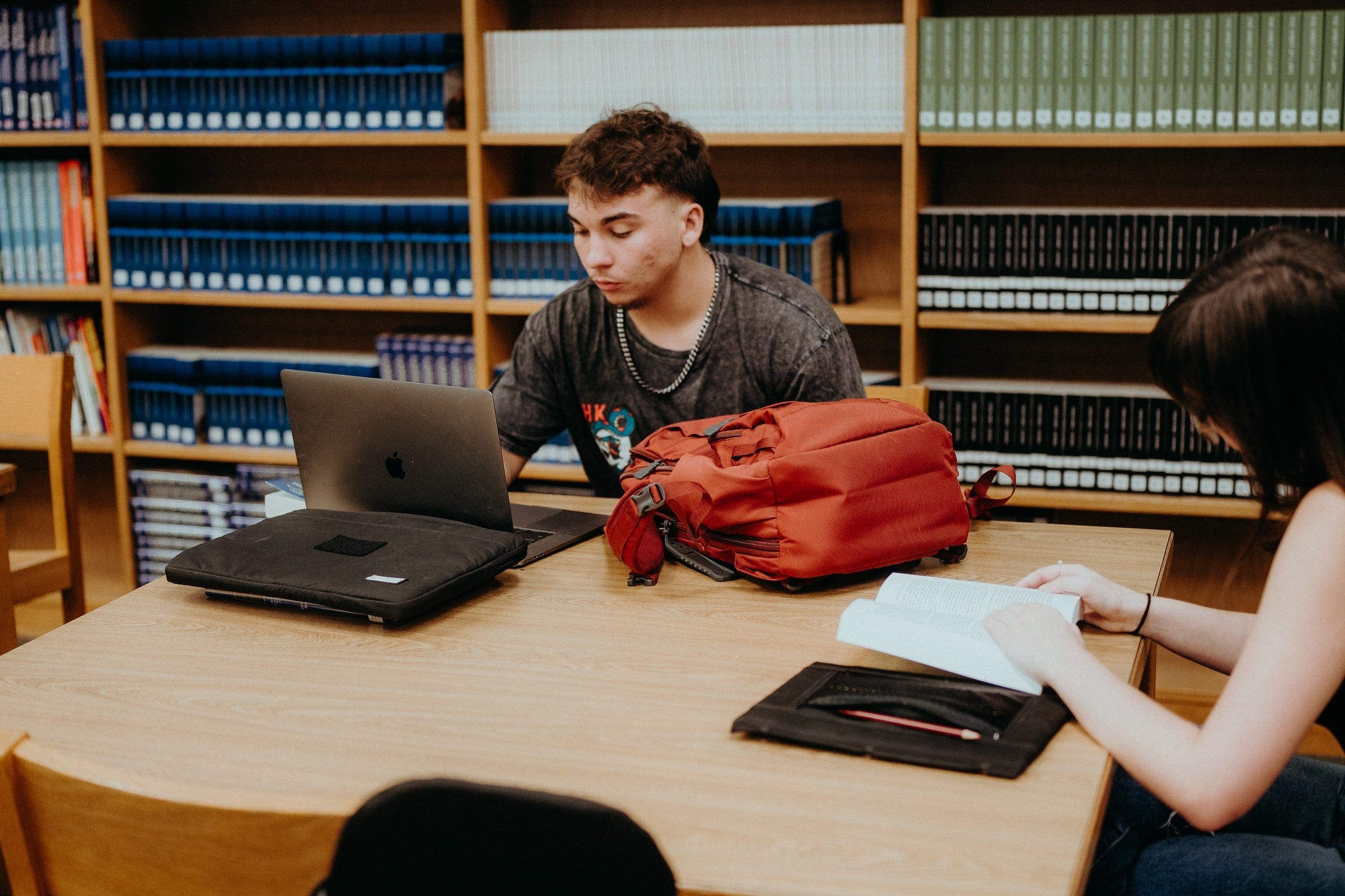 Two students sit at a library table. One is using a laptop, while the other is reading a book. A red backpack and a closed laptop are on the table, with bookshelves filled with volumes in the background.