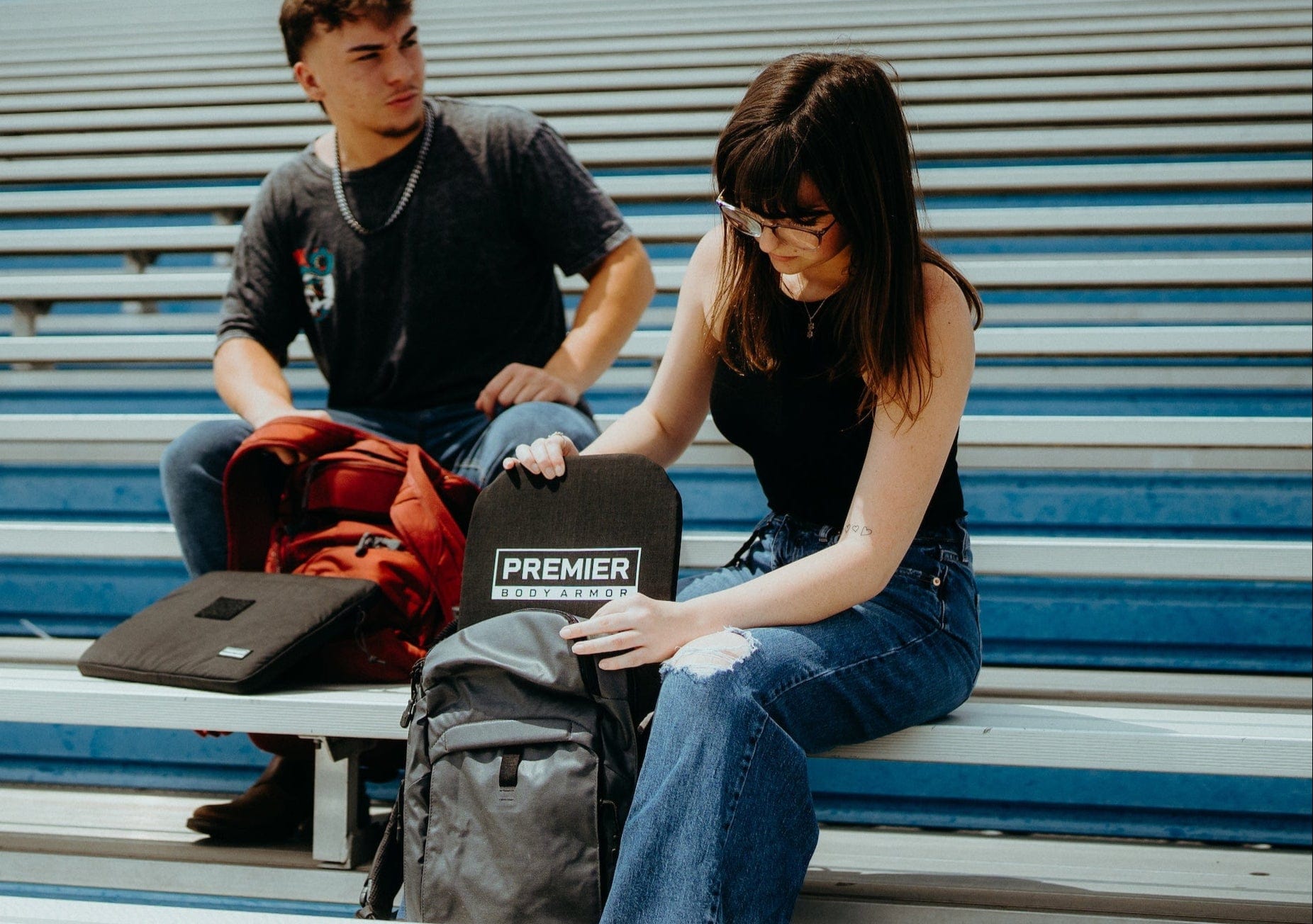 A young woman sitting on bleachers inserts a Premier Body Armor panel into her gray backpack, while a young man with a red backpack sits beside her. Both appear to be students.