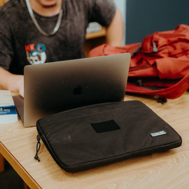 A person sits at a wooden table using a laptop. In the foreground, there's a closed Premier Body Armor Bulletproof Laptop Case with its sleek para-aramid ballistic core. Next to the laptop is an open red backpack, and a book is partially visible on the table.