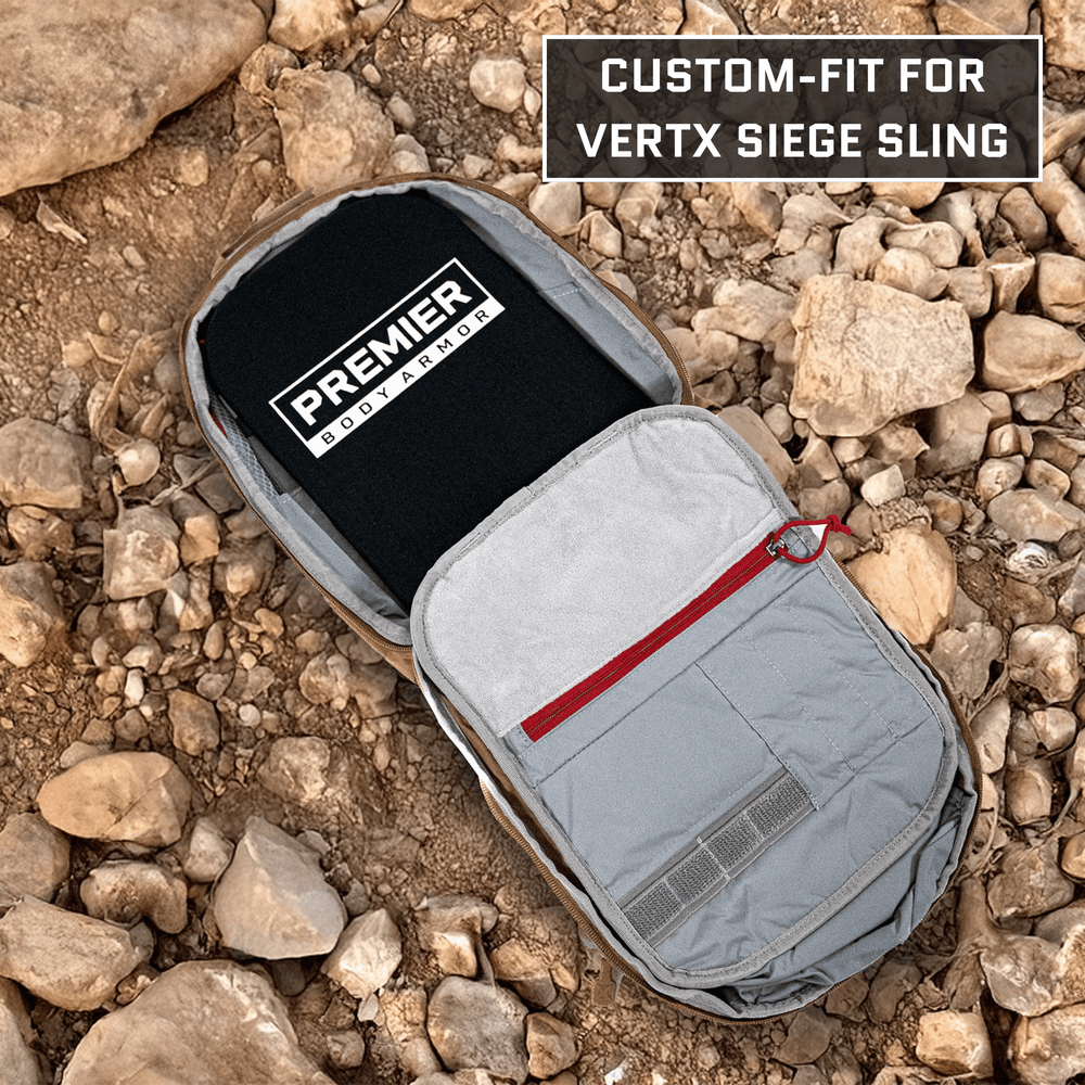 A gray backpack with a red zipper is open, showing the Premier Body Armor's Vertx Siege Sling Level IIIA/HG2 Backpack Insert. It rests on rocky ground, highlighting its custom fit and ultimate protection.