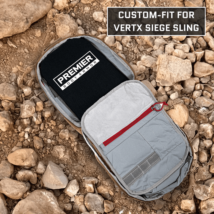 A gray backpack with a red zipper is open, showing the Premier Body Armor's Vertx Siege Sling Level IIIA/HG2 Backpack Insert. It rests on rocky ground, highlighting its custom fit and ultimate protection.