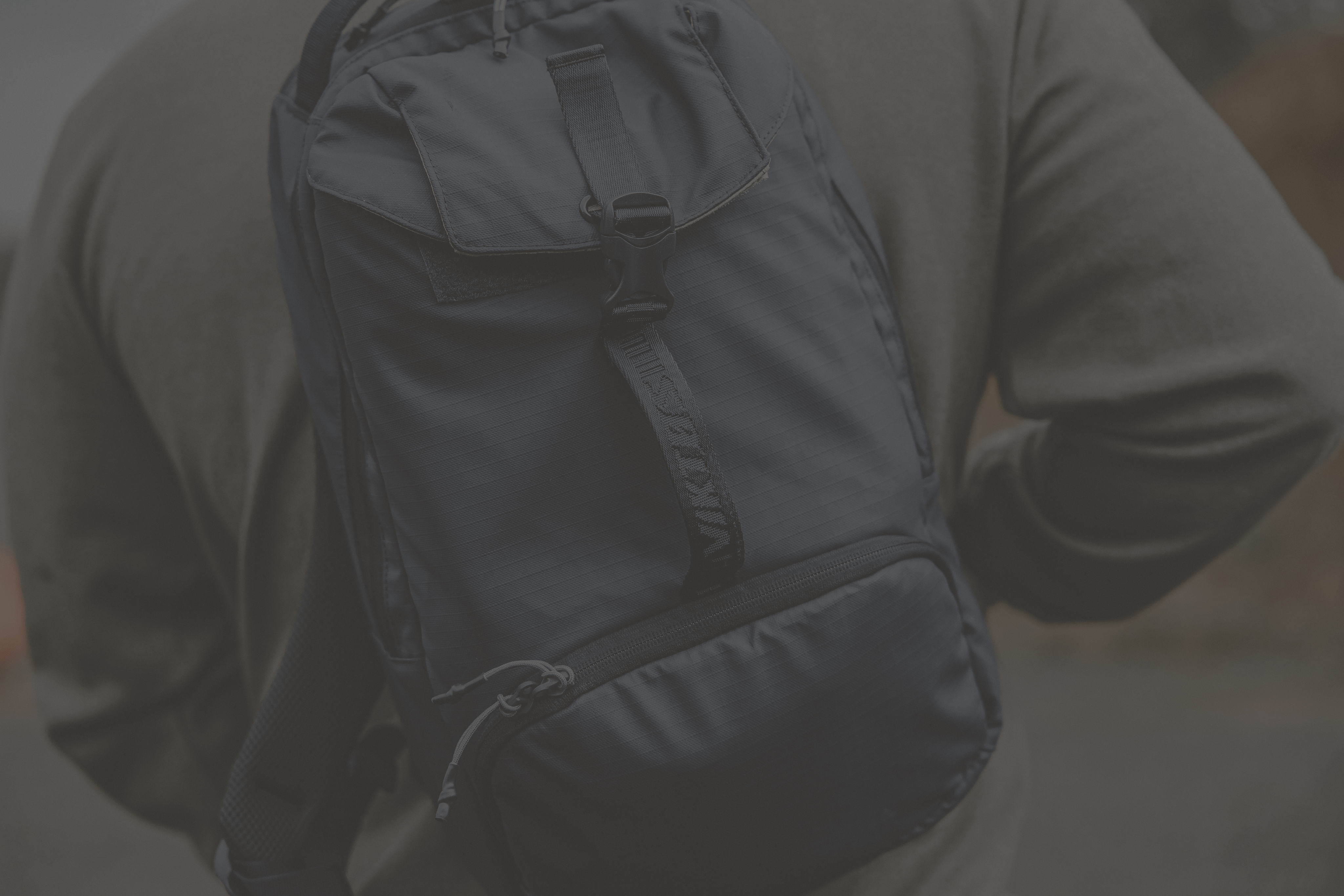 A person wearing a dark-colored backpack with multiple compartments and zippers. The image is slightly dim, and the background appears to be outdoors and blurred.