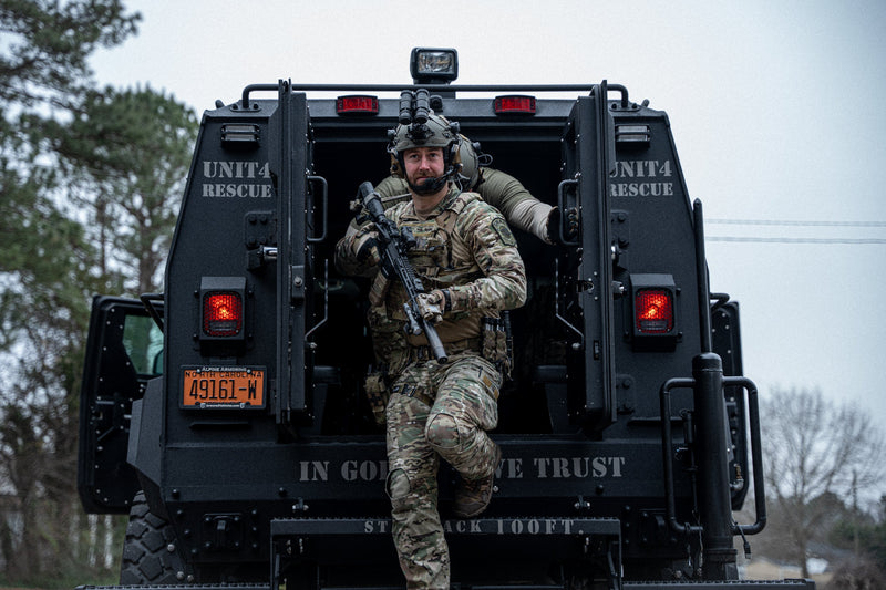 A uniformed armed officer steps out of the back of a black armored rescue vehicle labeled “UNIT 4 RESCUE” with open doors and emergency lights on, in an outdoor setting.