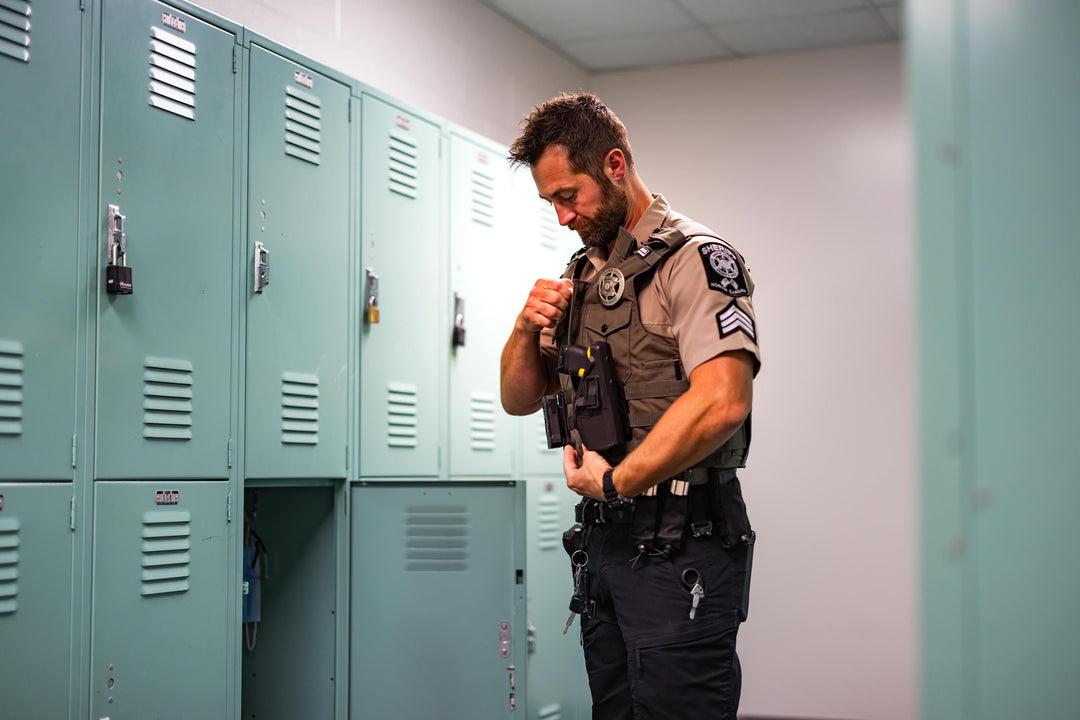 A police officer in uniform stands in a locker room, adjusting equipment on his vest in front of a row of light blue lockers. The setting appears quiet and well-lit.