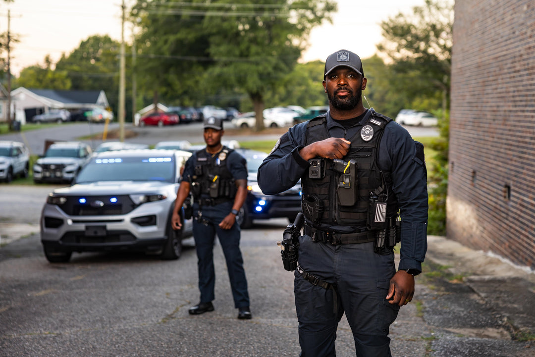 Two police officers stand outdoors in front of police vehicles with flashing lights; one officer stands in the foreground while the other stands farther back near the cars.