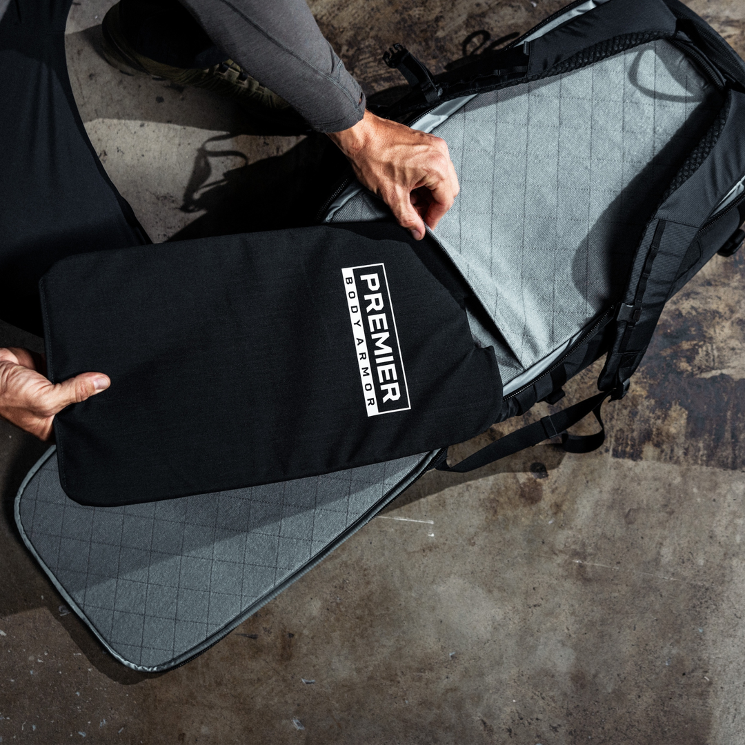 A person places a black Premier Body Armor panel into a gray, open backpack on a concrete floor. Only the person's hands and part of their legs are visible.
