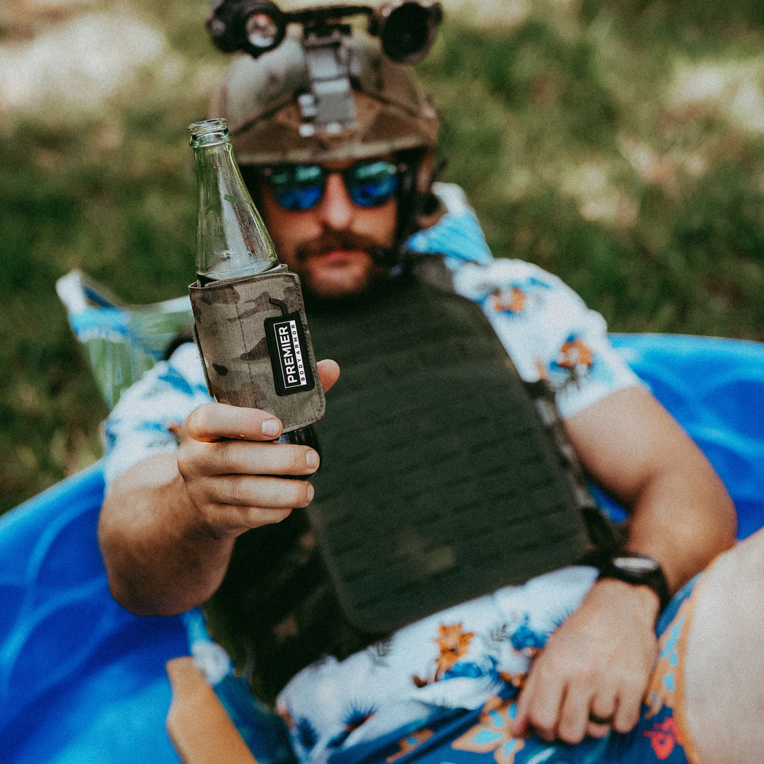 A person wearing a tactical vest, helmet with night vision goggles, and sunglasses sits in a blue kiddie pool, holding a bottle in a camouflage sleeve. The person is wearing a floral shirt and shorts.