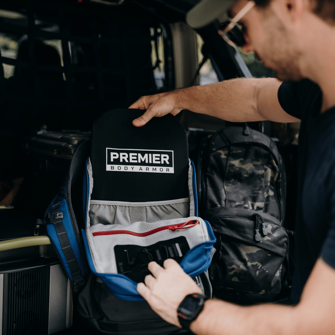 A person wearing a cap is placing a black armor plate labeled "Premier Body Armor" into a blue and gray backpack. Another camouflage backpack is visible in the background inside a vehicle.