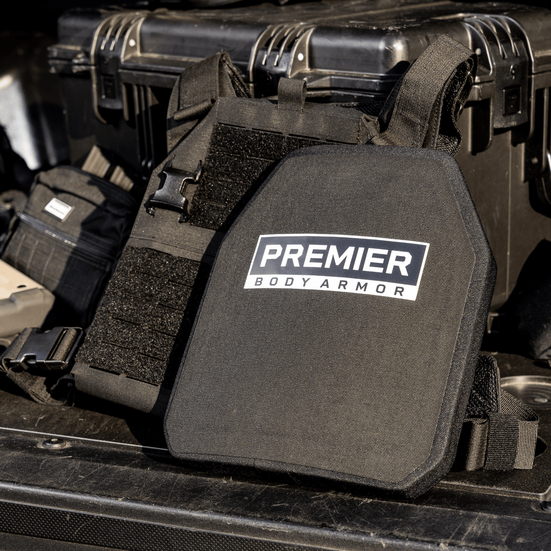 Body armor with a label reading "Premier Body Armor" is displayed in front of a stack of black tactical gear cases. The background includes various pieces of protective equipment.