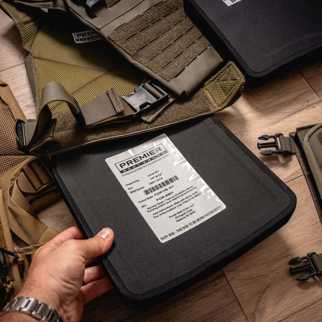 A hand holding a Premier Body Armor Fortis III+ ballistic plate (10"x12") with product label visible. A tan tactical vest and additional armor plates are visible in the background.