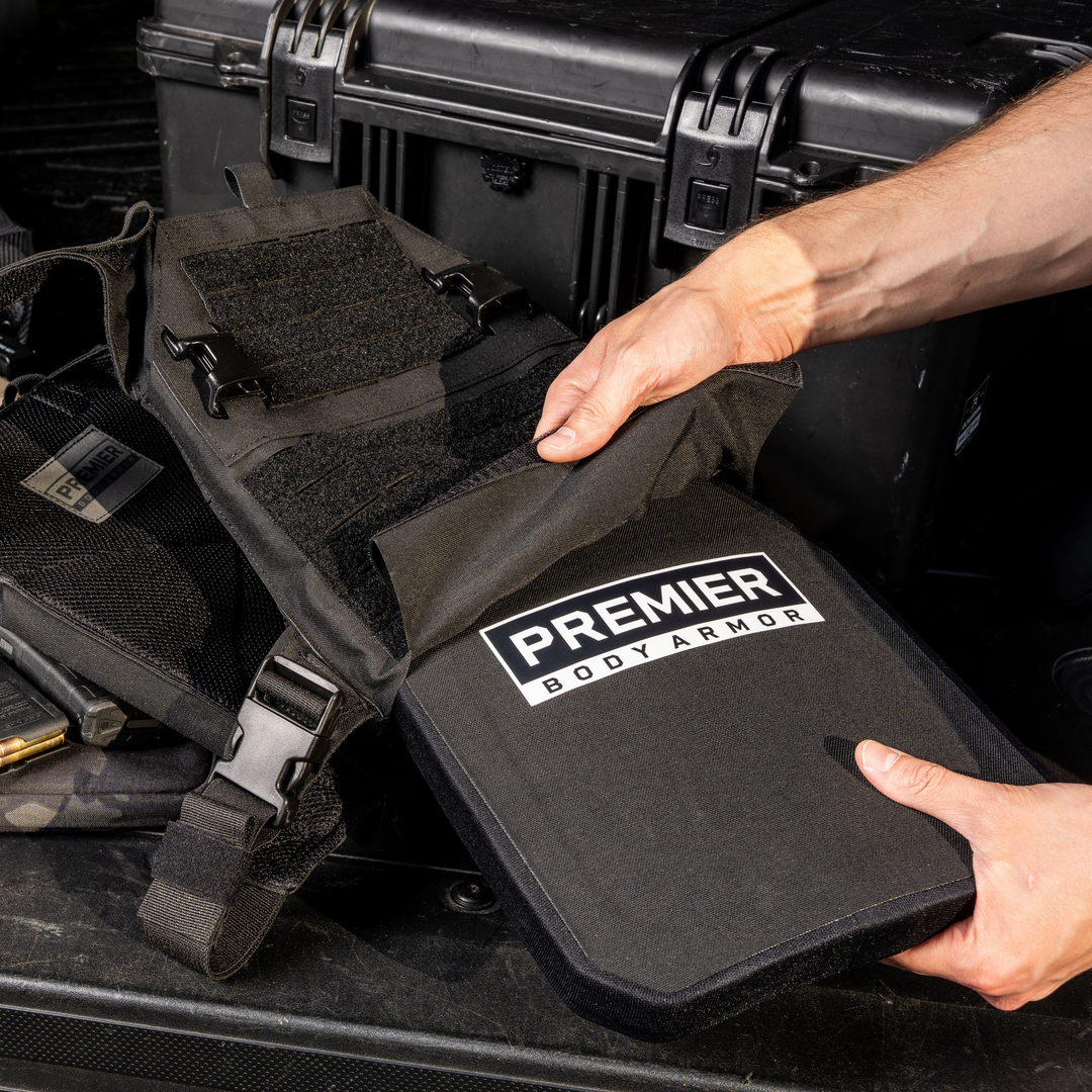 A person inserting a Premier Body Armor panel into a black tactical plate carrier, set in a vehicle trunk alongside a hard case and additional gear.