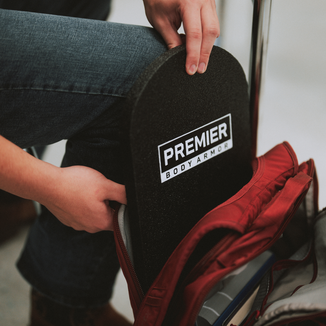 A person places a black Premier Body Armor plate into the main compartment of a red backpack while sitting down.