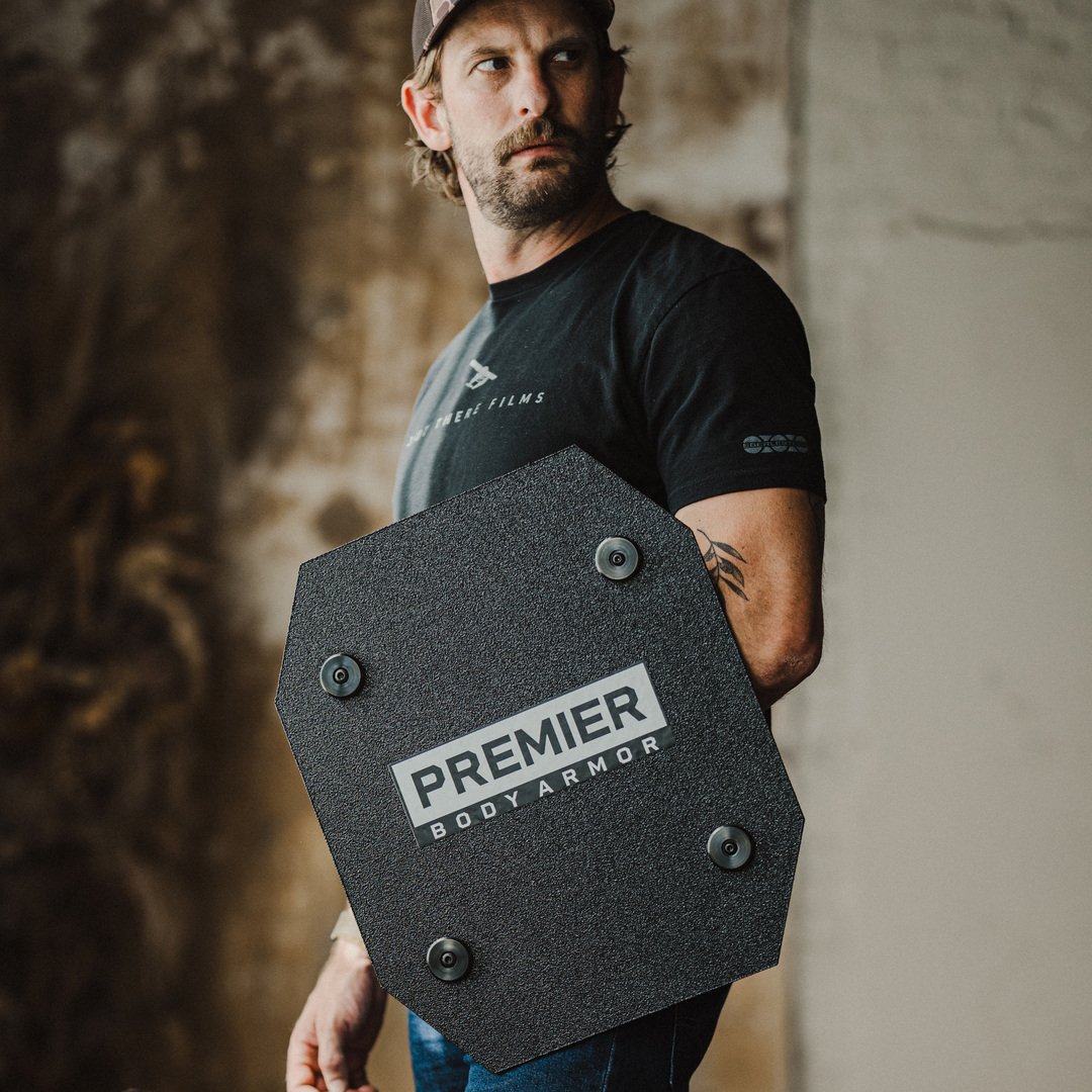 A man in a black t-shirt and cap holds a black, angular body armor shield labeled "Premier Body Armor" in front of a blurred indoor background.