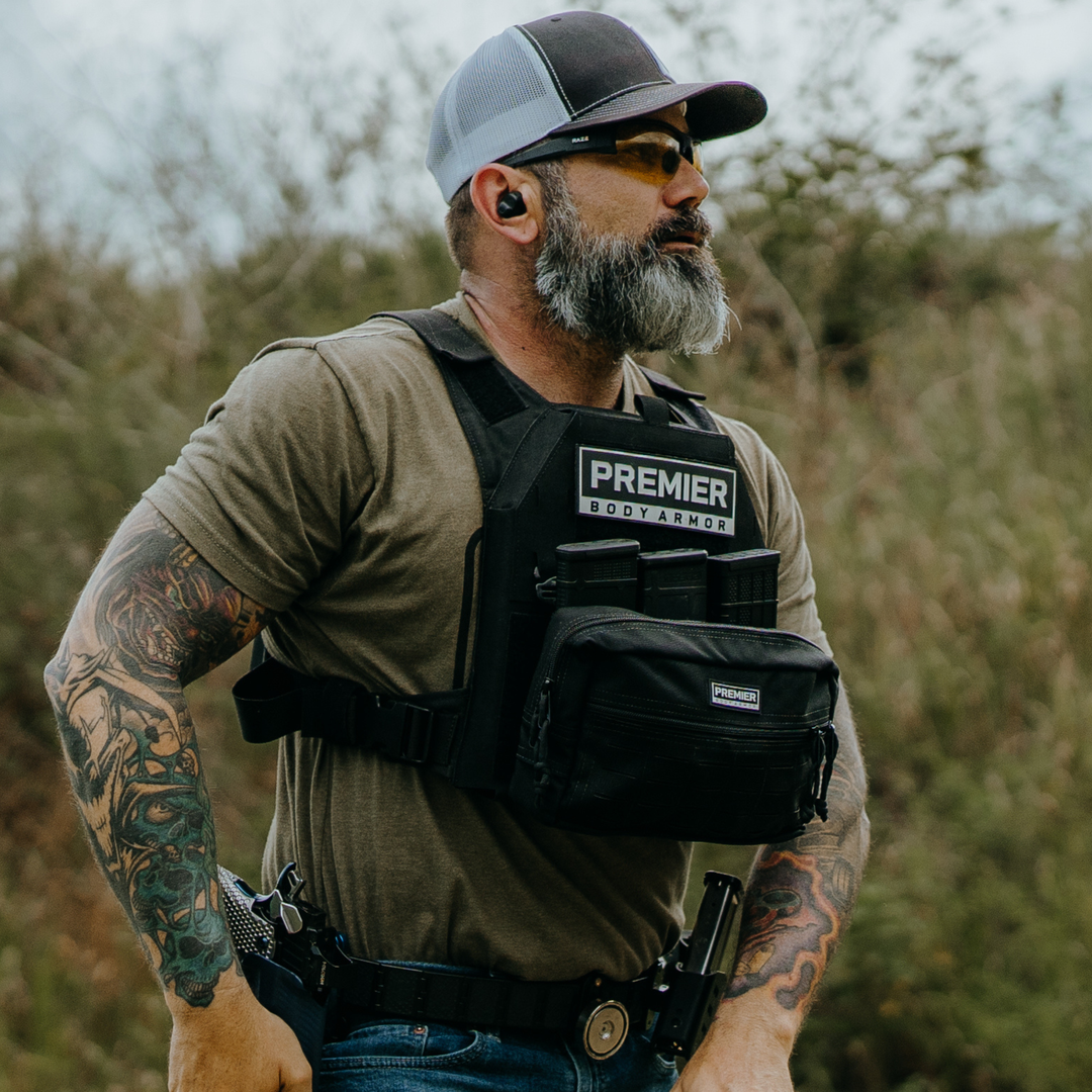 A bearded man wearing sunglasses, a cap, ear protection, and body armor stands outdoors. He has tattoos on his arms and carries a handgun in a holster and a pouch on his vest.