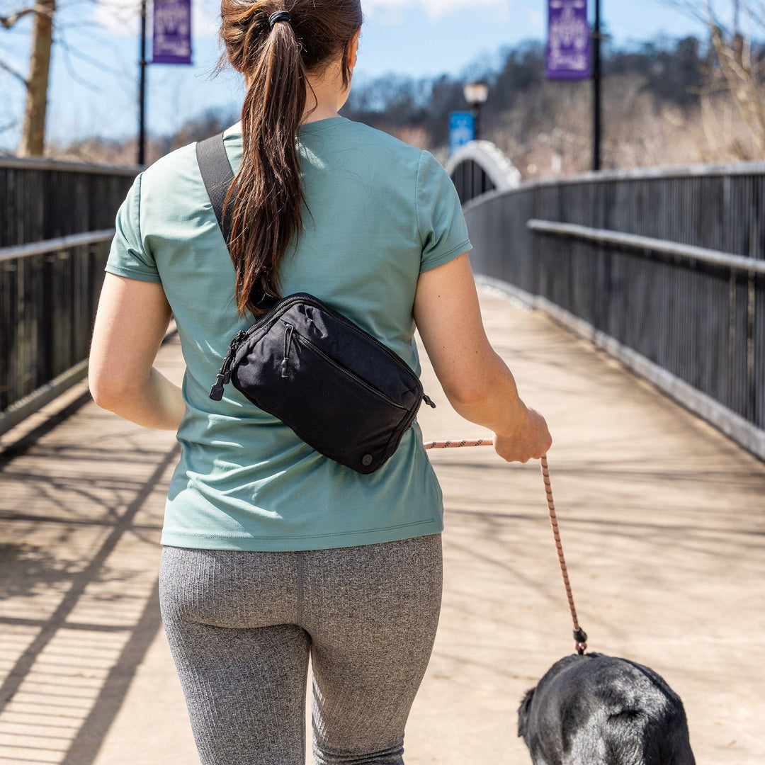 A woman with a ponytail walks on a sunlit path, her everyday companion—a black dog—by her side on a leash. She wears a green shirt, gray leggings, and carries the Vertx Everyday Fanny Pack across her back. Street lamps and trees line the path.