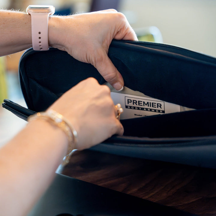 A person wearing a smartwatch is handling a Premier Body Armor Bulletproof Laptop Case with Level IIIA Protection from their bag's zippered compartment, with the hands and case in focus against a blurred background.