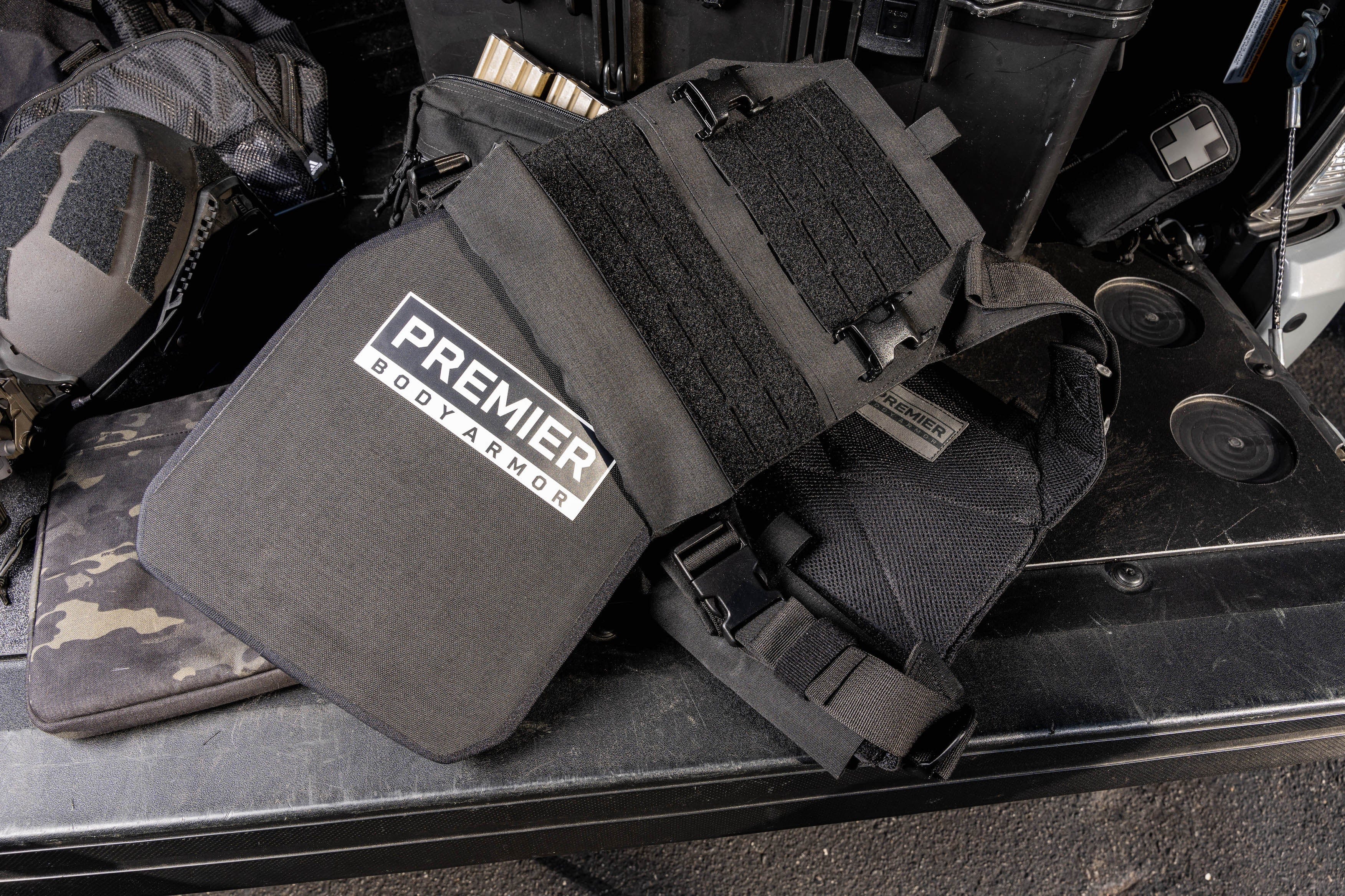 A piece of body armor from Premier Body Armor is displayed on a table, featuring a black protective panel with straps and a Velcro attachment area. Designed for tactical professionals, the Fortis Level III+ GT Loadout (old) offers advanced ballistic protection. Various other tactical gear items are visible in the background.