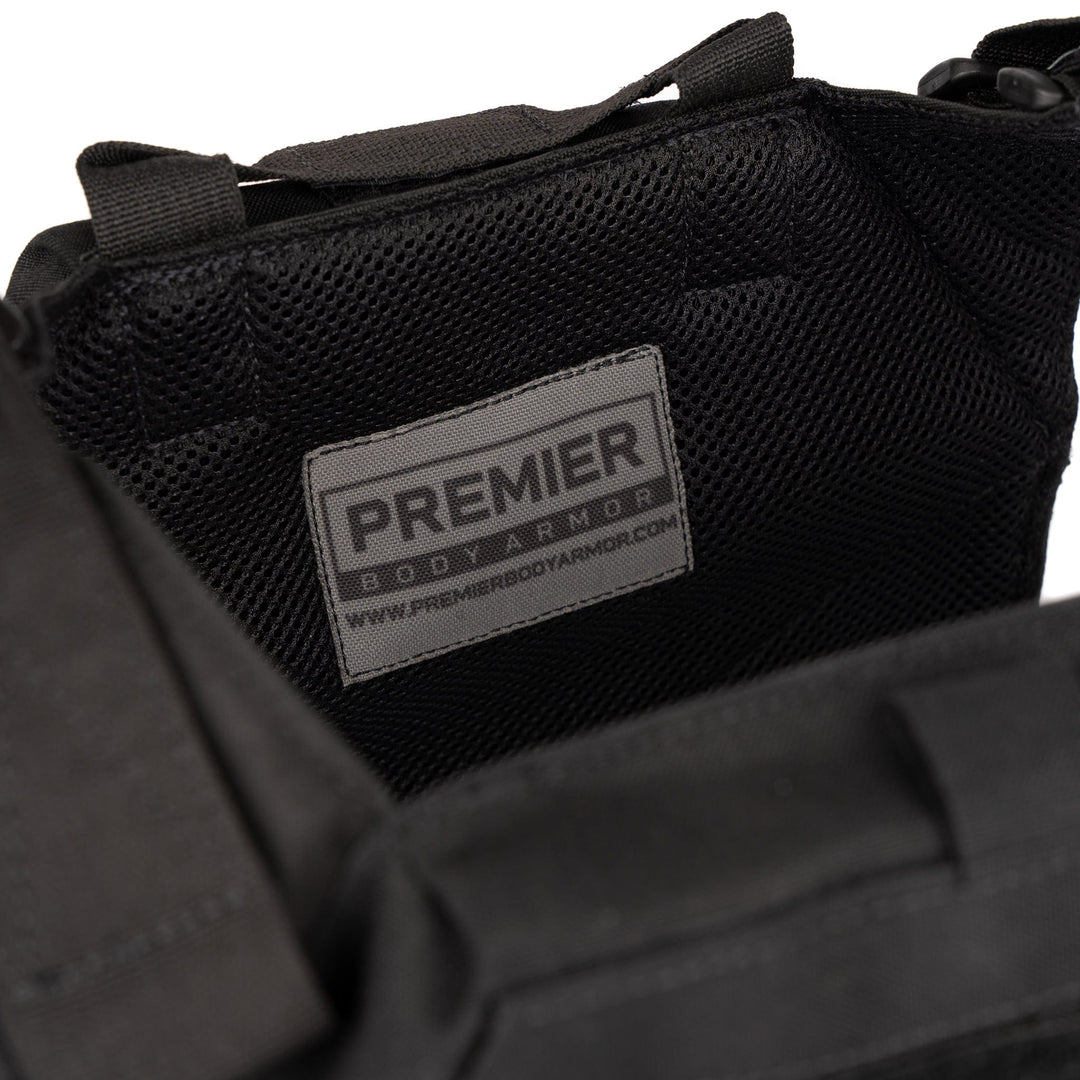 A close-up of the Premier Body Armor Fortis Alpha Carrier, showcasing its textured black surface. The label on the armor reads "Premier Body Armor" and includes the website "www.premierbodyarmor.com." Black straps or loops are visible around the armor for securing it. The Fortis Alpha Carrier is crafted from durable 500D Nylon and features laser-cut MOLLE.