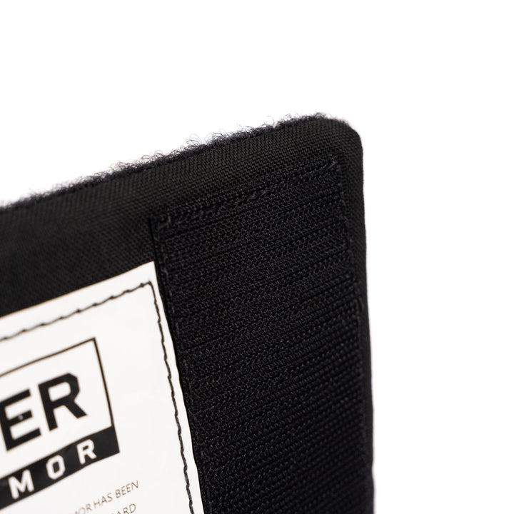 Close-up of a black fabric item showing a corner seam with detailed stitching, indicative of its high-quality craftsmanship. A partially visible white label with black text is attached to the item. The background is white. This could be a component of the Premier Body Armor's SOE FUPA Level IIIA Insert for added protection.