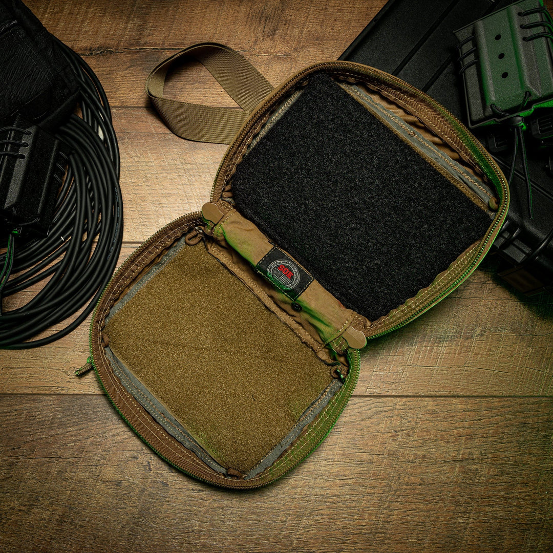 An SOE FUPA Level IIIA Insert by Premier Body Armor, featuring an interior lined with hook-and-loop (Velcro) panels, lies open on a wooden surface surrounded by coiled cables and other tactical equipment, such as a black case, green-stuffed pouches, and an SOE Fanny Pack. A strap is attached to the insert.